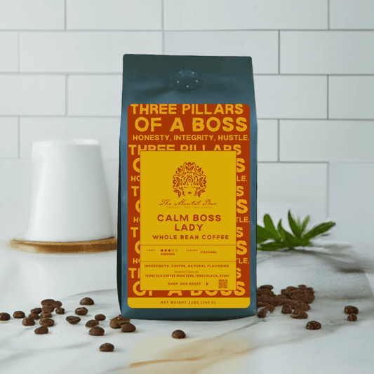 A bag of 'Calm Boss Lady' whole bean coffee with a yellow and black label, placed on a white surface with a ceramic cup and coffee beans scattered around.
