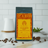 A bag of 'Calm Boss Lady' whole bean coffee with a yellow and black label, placed on a white surface with a ceramic cup and coffee beans scattered around.