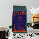 A pack of 'Rhythm & Riches' coffee beans with a medium roast level, sourced from Indonesia and India. The packaging is navy blue with orange accents and text, and there is a cup of coffee and coffee beans on the foreground.