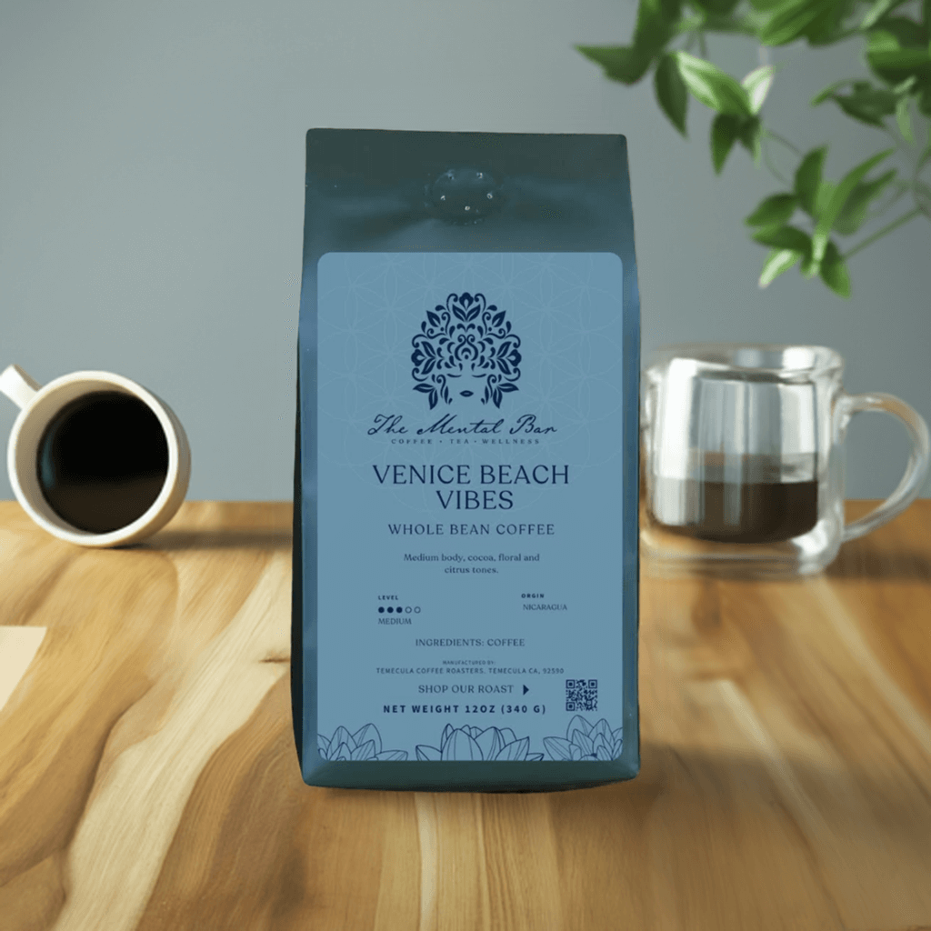 A pack of 'The Mental Bar' Venice Beach Whole Bean Coffee, medium roast, with a graphic design on the packaging, placed on a wooden table with a cup of coffee and a plant in the background.