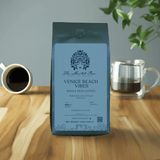 A pack of 'The Mental Bar' Venice Beach Whole Bean Coffee, medium roast, with a graphic design on the packaging, placed on a wooden table with a cup of coffee and a plant in the background.